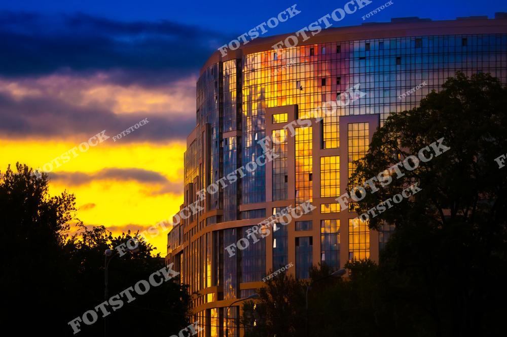 На фотографии изображено современное здание в Санкт-Петербурге, фасад которого отражает яркие краски заката. Закатное небо окрашено в теплые желтые и оранжевые тона, контрастируя с темными силуэтами деревьев и урбанистическими линиями здания. Это сочетание природы и архитектуры создает эффектную композицию, подчеркивающую гармонию между городом и его окружением.